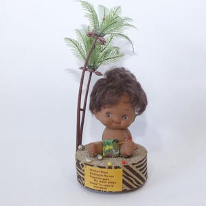 May include: A vintage doll with dark hair and a smiling face sits atop a decorative base with a palm tree. The base has a brown and tan zebra stripe pattern and text. Small pearls surround the base.