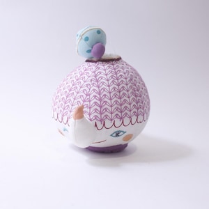May include: A whimsical, handmade plush doll with a round body and a small, hat-like detail on top. The doll's body is patterned with a purple and white design. It has a white face with blue eyes and peach-colored cheeks.