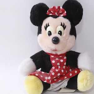 K&ouml;nnte beinhalten: Ein Pl&uuml;schtier aus schwarzem und wei&szlig;em Mickey Mouse, das ein rotes Kleid mit wei&szlig;en Punkten und einer roten Schleife tr&auml;gt. Das Spielzeug hat gro&szlig;e schwarze Augen und eine schwarze Nase.