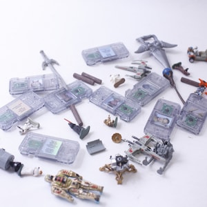 Könnte beinhalten: Eine Sammlung kleiner Spielzeuge aus Plastik, darunter ein Raumschiff, ein Roboter, ein Schwert und ein paar weitere Figuren. Die Spielzeuge sind auf einer weißen Oberfläche verstreut.