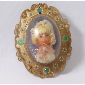 May include: A vintage doll encased in a gold-colored, ornate frame. The doll has blonde hair, blue eyes, and wears a white dress and bonnet. The frame is decorated with green and white accents, and the doll is set against a purple background.