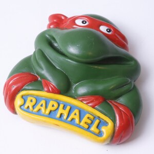 May include: Green Teenage Mutant Ninja Turtles character Raphael figurine with a red bandana and a yellow sign that says "Raphael".