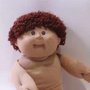 Op de afbeelding: Een vintage pop met een lichte huidskleur, bruin garen haar en grote, expressieve ogen. De pop heeft een zacht lichaam en draagt geen kleding. De armen van de pop zijn uitgestrekt en ze heeft een lichte glimlach. Een klassiek speelgoed.
