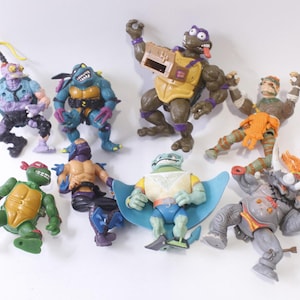 May include: A collection of nine Teenage Mutant Ninja Turtles action figures, including Leonardo, Donatello, Raphael, Michelangelo, Shredder, Bebop, Rocksteady, and two other figures. The figures are made of plastic and are painted in various colors, including green, blue, red, orange, and purple.