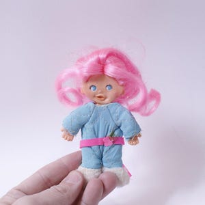 Puede incluir: Una pequeña muñeca con el pelo rosa que lleva un mono acolchado azul y blanco con una faja rosa y un adorno floral. La muñeca tiene ojos azules y una sonrisa pintada.