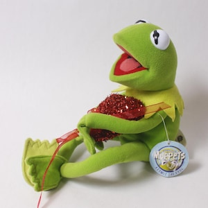 以下が含まれることがあります： 緑色のカエルのカーミットのぬいぐるみ。座って赤いハート型のオブジェクトを持っています。カエルは黄色の襟と開いた口をしています。マペットのロゴが付いたタグが付いています。