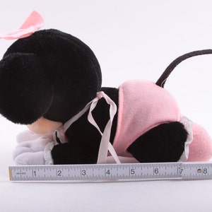 May include: A black and pink plush toy of a cartoon mouse wearing a pink dress and a pink bow. The toy is lying on its stomach with its head turned to the side. The toy is about 7 inches long.
