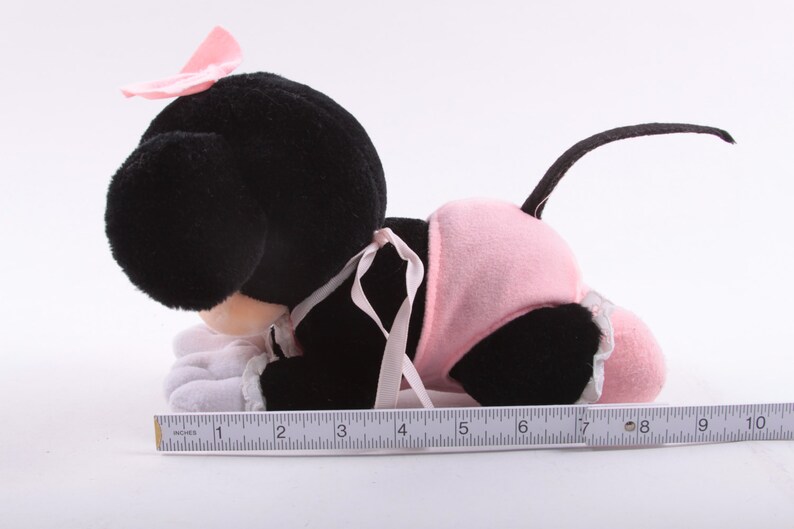 May include: A black and pink plush toy of a cartoon mouse wearing a pink dress and a pink bow. The toy is lying on its stomach with its head turned to the side. The toy is about 7 inches long.