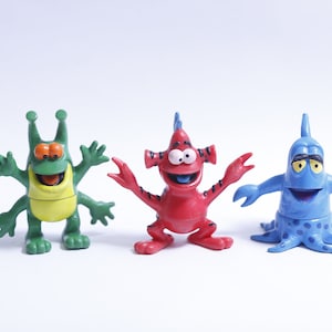 May include: Three colorful cartoon monster figurines. The first is green with yellow accents and four arms. The second is red with black stripes and two arms. The third is blue with white spots and eight arms.