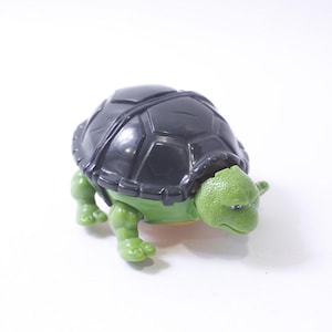 May include: A green and black plastic toy turtle with a hard shell. The turtle has a cartoon-like face with a closed mouth and small eyes.