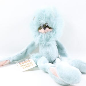 May include: A light blue plush toy with long limbs and a fluffy mane surrounding a face with a dark green mask, a stitched nose, and a tag that reads BAMAR ORIGINAL SAMPLE. The toy features soft, textured fur and light pink accents on the paws.