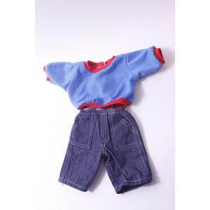May include: A blue and red doll outfit, consisting of a short-sleeved top and denim shorts. The top is light blue with red trim around the neck and sleeves. The shorts are dark blue denim with white stitching and pockets.