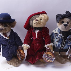 May include: Three plush teddy bears dressed in distinct outfits. The left bear wears a blue pinstripe suit, hat, and glasses. The center bear wears a red velvet dress and hat. The right bear wears a denim jacket and black headpiece.