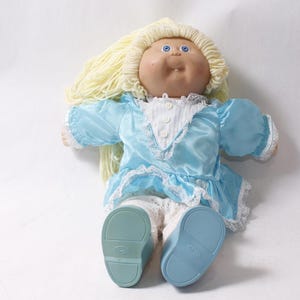 May include: A doll with light yellow yarn hair, blue eyes, and a round face. The doll wears a shiny light blue dress with white lace trim and a white collar, paired with white lace-trimmed pants and solid light blue shoes with flat soles.