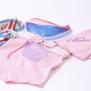 Könnte beinhalten: Eine Sammlung von Babyartikeln in Rosa, Weiß und Blau. Enthält einen rosa Strampler mit einem lila Aufnäher, ein rosa Handtuch, eine blau-rosa Federtasche und weiße Turnschuhe mit roten und blauen Akzenten. Alle Artikel tragen den Text "L.A. Gear".