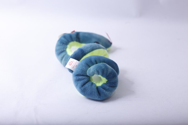 Pode incluir: Um brinquedo de pel&uacute;cia azul e verde em forma de cobra. A cobra est&aacute; enrolada em espiral. O brinquedo tem uma pequena etiqueta vermelha anexada.