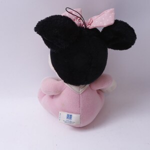 May include: A plush Minnie Mouse doll, viewed from the back, with black ears and head, a pink bow with white polka dots, and a pink dress. The doll has a white face and is sitting down.