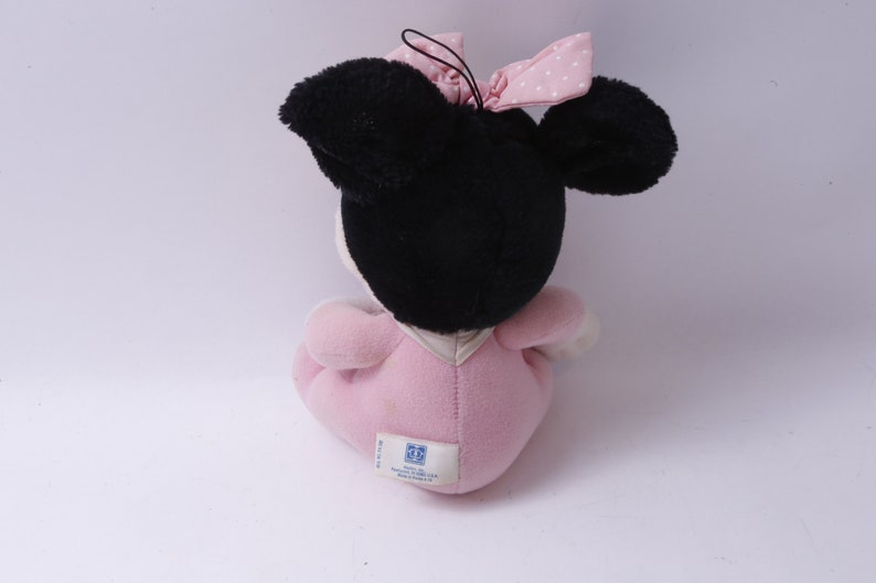 May include: A plush Minnie Mouse doll, viewed from the back, with black ears and head, a pink bow with white polka dots, and a pink dress. The doll has a white face and is sitting down.