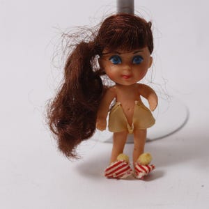 May include: A small vintage doll with brown hair in a ponytail and blue eyes. The doll is wearing a yellow skirt and red and white striped shoes. The doll is standing on a white surface.