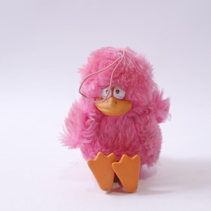 May include: A pink plush toy shaped like a bird with a sad expression. The toy has orange feet and a wire loop on its head.