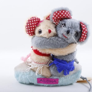 Puede incluir: Dos ratones de peluche abrazándose. Uno es beige con un corazón rojo y orejas de lunares rojos. El otro es gris con orejas de lunares rojos. Están sobre una base de nube azul con la palabra "ALOHA" en una etiqueta rosa.