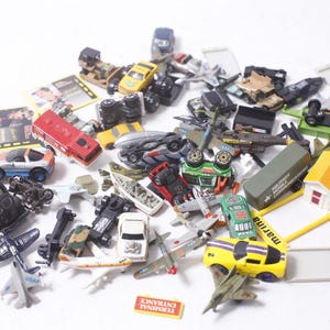 May include: A collection of miniature toy vehicles, including cars, trucks, airplanes, and military vehicles. The toys are in various colors, such as red, yellow, green, and black. Some toys are upside down. A small sign reads "TERMINAL ENTRANCE."