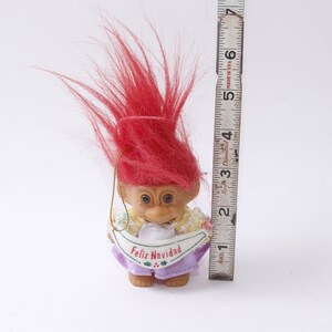 May include: A red-haired troll doll wearing a purple dress with a white banner that says "Feliz Navidad".