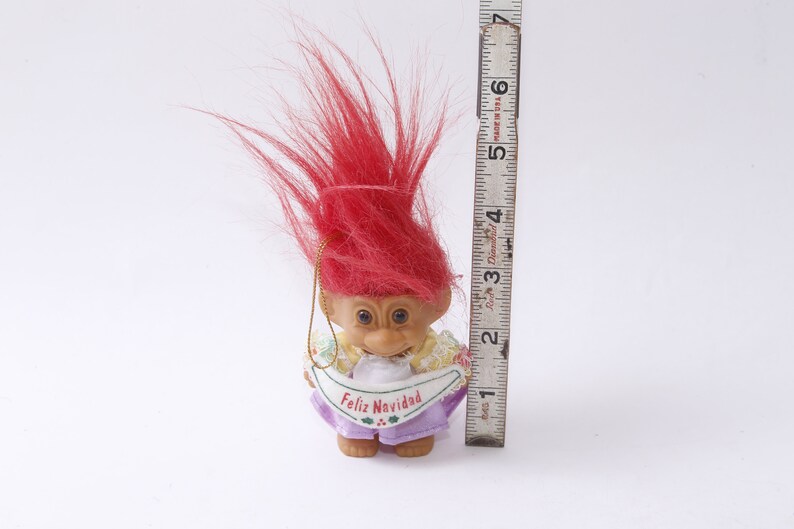 May include: A red-haired troll doll wearing a purple dress with a white banner that says "Feliz Navidad".