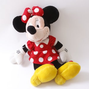 May include: A plush toy of Minnie Mouse wearing a red dress with white polka dots and a red bow with white polka dots. She has black ears and a black nose. Her shoes are yellow.