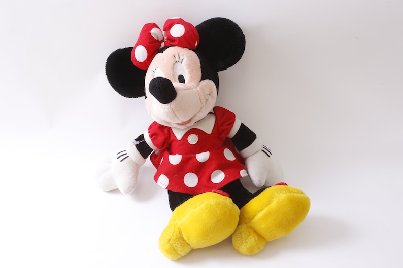 May include: A plush toy of Minnie Mouse wearing a red dress with white polka dots and a red bow with white polka dots. She has black ears and a black nose. Her shoes are yellow.