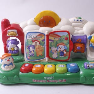 May include: A VTech Discovery Nursery Farm toy with a green base, colorful buttons, and animal figures. The toy features a red barn, a yellow sun, and an open book with illustrations. The toy has the text "Old MacDonald Had a Farm" and "E-I-E-I-O".