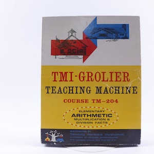 May include: A vintage TMI-Grolier Teaching Machine box. The box has a yellow and white color scheme with red and blue graphics of buildings. Text on the box includes "Teaching Machine" and "Elementary Arithmetic Multiplication & Division Facts."