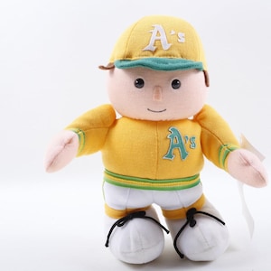 以下が含まれることがあります： オークランド・アスレチックスの「A's」ロゴが入った黄色い野球のユニフォームを着たぬいぐるみ。人形は「A's」ロゴ入りの黄色い帽子、黄色のシャツ、白いショートパンツ、黒い靴紐の白い靴を履いています。人形は腕を広げて立っています。