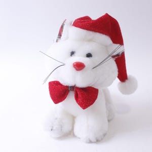 May include: White plush cat wearing a red Santa hat with a white pom-pom and a red bow tie.