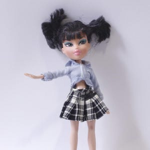 Puede incluir: Una falda de cuadros negros y blancos con una camisa azul abotonada. La muñeca tiene el pelo negro con flequillo y ojos azules.