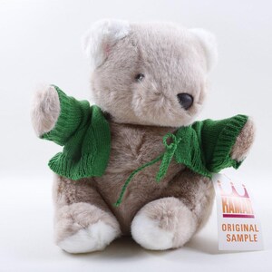 May include: A plush teddy bear with light brown fur, white accents on the ears and feet, and a dark nose. The bear wears a green knitted cardigan with a tie closure. A white tag attached to the arm features the text KAMAI ORIGINAL SAMPLE.