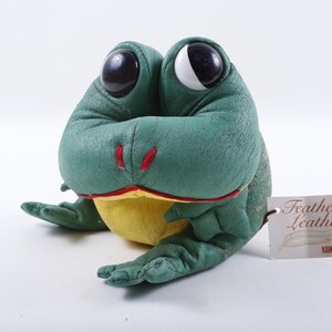 May include: Green plush frog with large black eyes and a yellow belly. The frog is sitting on a white surface with a tag that reads "Feather Leather" and "KAMAR."