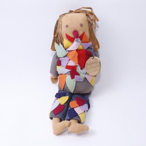May include: A handmade rag doll scarecrow with a gray shirt and blue pants. The doll is decorated with colorful fabric scraps and has a straw-like hair. The doll has a tag that reads "Good journey with Kenneth & Sandra".