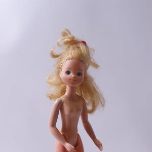 May include: A plastic doll with blonde hair and blue eyes. The doll is wearing no clothes and has a simple, classic design.