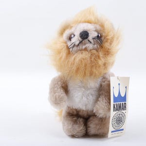 May include: A vintage plush lion toy with a light brown mane and body. The toy has a black nose and whiskers, and a tag that reads "KAMAR" with a crown and a compass rose design. The toy is standing upright.