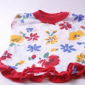Puede incluir: Top blanco con dobladillo y cuello con volantes rojos, adornado con coloridos estampados florales. Las flores son en tonos rojos, amarillos y azules. La parte superior probablemente está hecha de una tela suave y ligera.