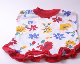 May include: White top with a red ruffled hem and neckline, adorned with colorful floral patterns. The flowers are in shades of red, yellow, and blue. The top is likely made of a soft, lightweight fabric.