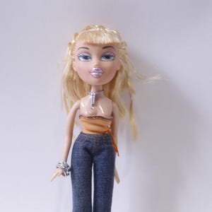 May include: A blonde doll with blue eyes and a light orange top. She is wearing blue jeans and a silver necklace.