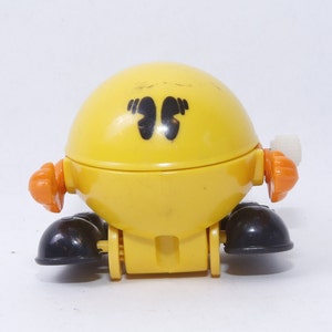 May include: A yellow wind-up toy shaped like a Pac-Man character with black feet and orange arms. The toy has a white winder on the side.