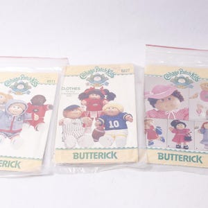 May include: Three vintage Butterick sewing patterns for Cabbage Patch Kids clothing. The patterns are in plastic sleeves and show illustrations of the dolls in different outfits. The patterns include the text "CLOTHES" and "BUTTERICK".