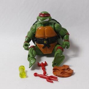 May include: A green Teenage Mutant Ninja Turtles action figure with a red eye mask, orange chest plate, and black belt featuring an R emblem. The figure is accompanied by two red trident-like weapons, a brown circular accessory, and a translucent yellow cylinder.
