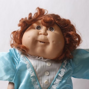May include: A doll with auburn hair and a round face featuring dimples and brown eyes. The doll wears a bright blue satin dress with a white pinstriped bodice, white lace trim along the collar and sleeves, and three small white buttons.