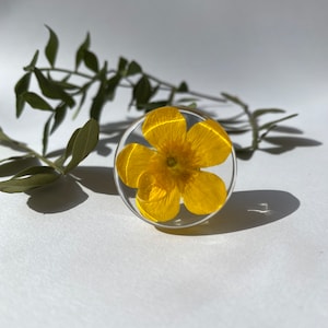 May include: A clear resin ring featuring a vibrant yellow flower. The flower is encased in a circular, transparent resin setting. Green leaves are in the background. The ring is set against a white background.