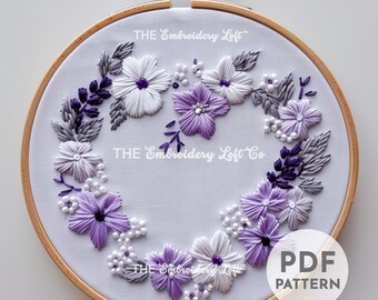 May include: A white embroidery hoop with a purple and white floral design. The design is in the shape of a heart and includes small white beads. The text "THE Embroidery Loft Co" is visible in the center of the hoop. The text "PDF PATTERN" is visible in the lower right corner of the hoop.