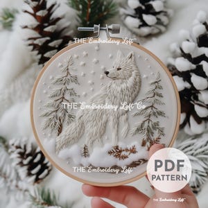 Könnte beinhalten: Stickmuster eines weißen Wolfes in einem Holzreifen mit einem schneebedeckten Wald im Hintergrund. Das Muster enthält einen Wolf, Kiefern und Schneeflocken. Der Text "THE Embroidery Loft Co" ist auf dem Reifen sichtbar.
